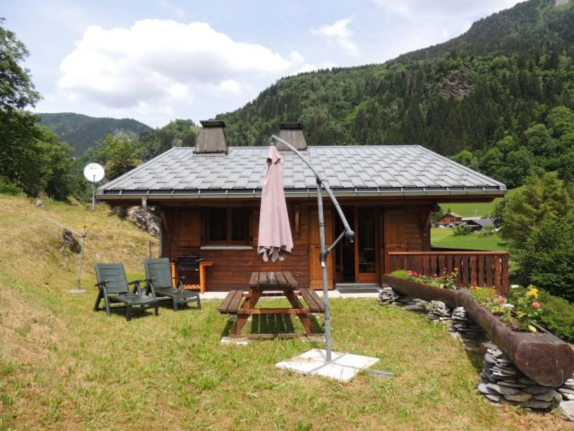 Terrasse estivale tout en herbe pour profiter du soleil de Haute-Savoie. Table de jardin avec bancs, parasol , chaises et deux bains soleils sont à votre disposition.