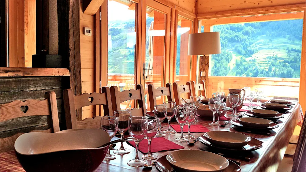 Grande table intérieure dressée pour un repas de 12 personnes au Chalet B à Vars, installée devant une grande fenêtre ouverte sur la montagne.