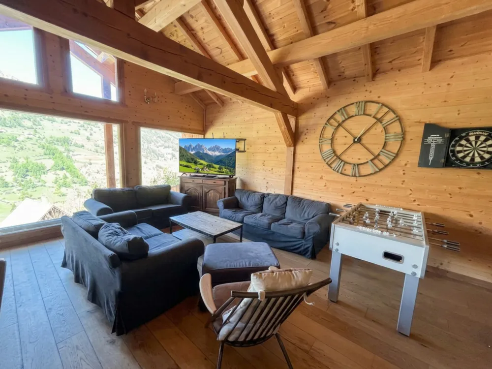 Grand salon du Chalet B à Vars avec canapé en U, TV écran plat, baby-foot, jeu de fléchettes et trois grandes baies vitrées s'ouvrant sur la montagne.