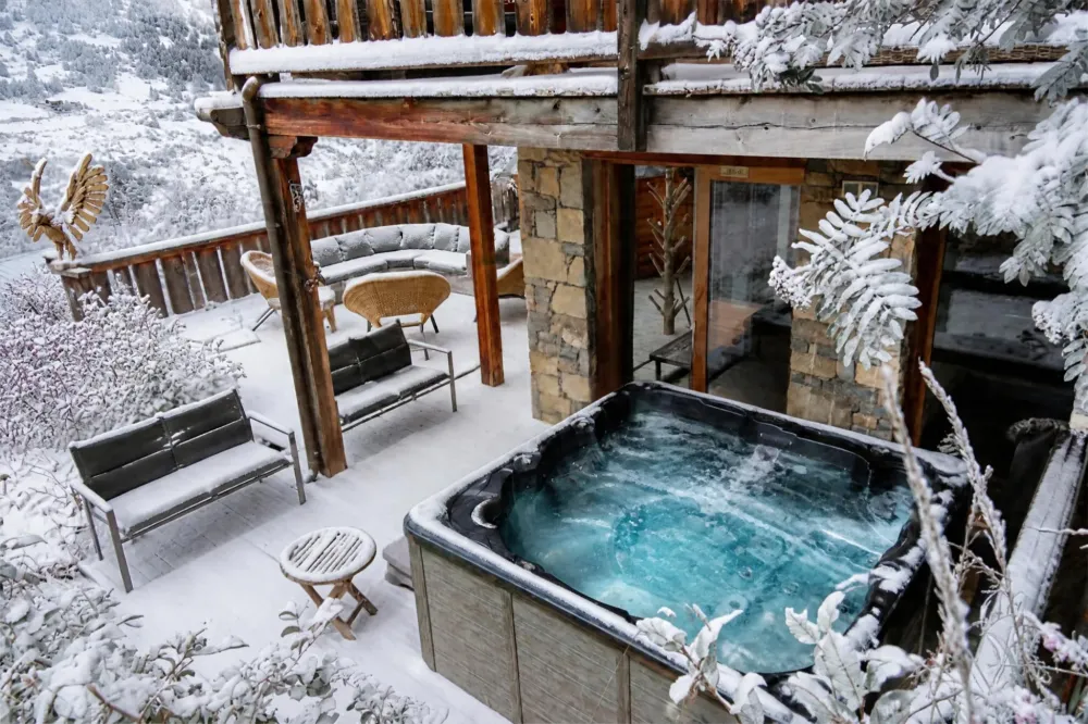 The snow-covered terrace of Chalet D in Vars, featuring an outdoor hot tub with turquoise water, snow-covered benches, patio furniture, and rattan armchairs, a wooden eagle sculpture, frost-covered shrubs, and the white valley in the background