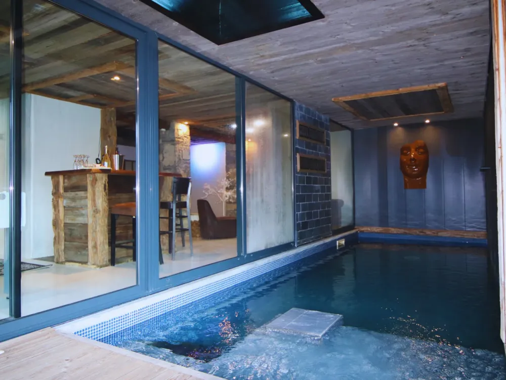 Piscine intérieure du Chalet D à Vars, bassin à mosaïque bleue, plafond en vieux bois, sculpture de visage au mur, grandes baies vitrées ouvrant sur un espace bar en bois brut avec tabourets et fauteuil en cuir