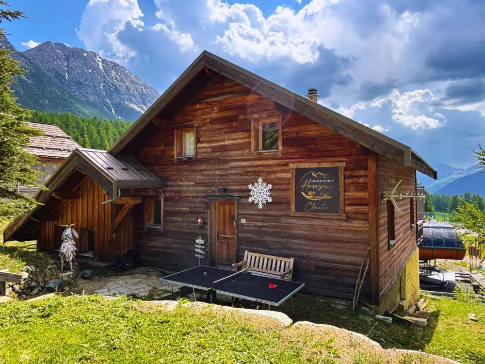 Façade du chalet La Clautre à Montgenèvre en été avec table de ping-pong et la gare du télémix des Chalmettes en arrière-plan.