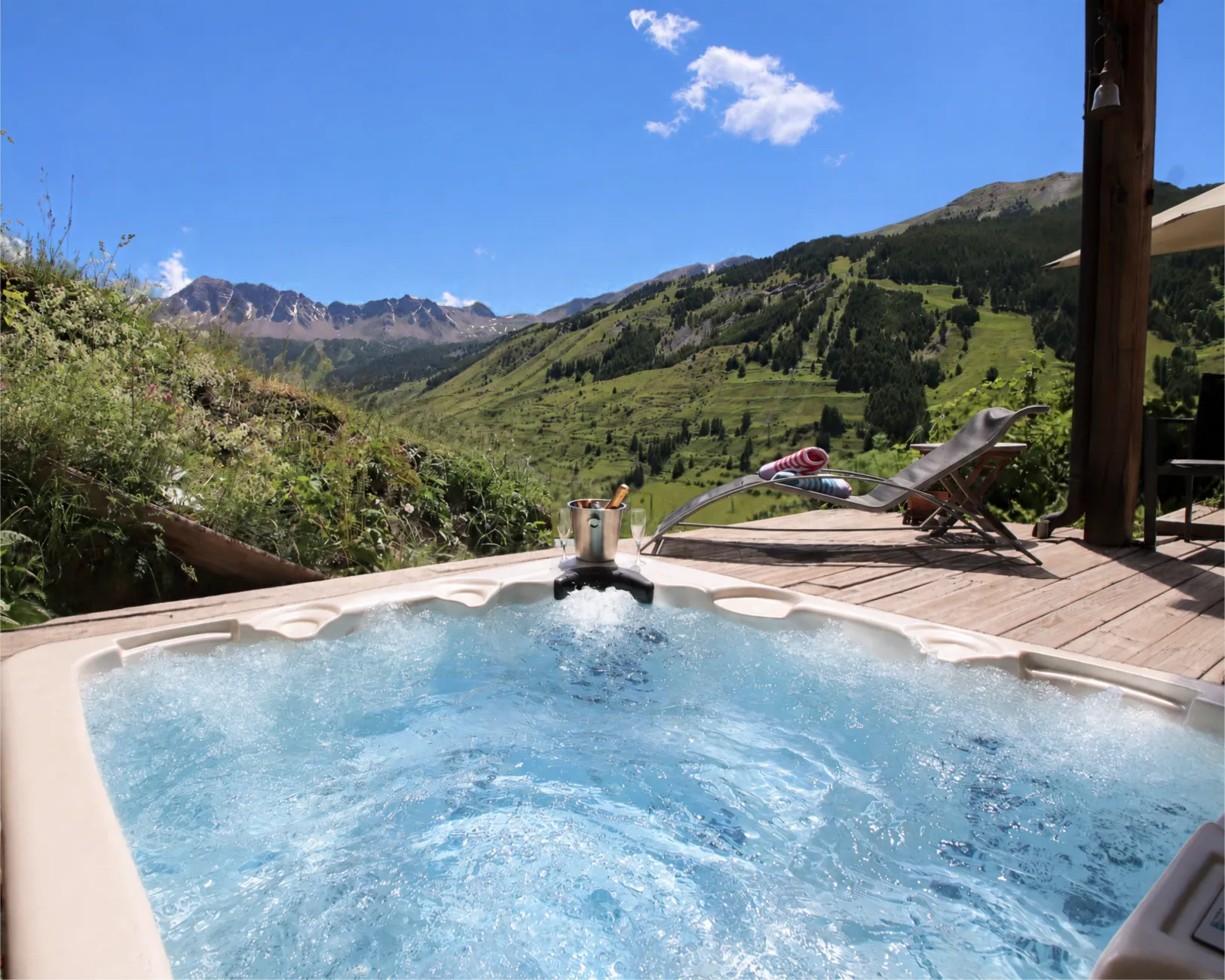 Jacuzzi extérieur bouillonnant du Chalet D à Vars avec seau à champagne et flûtes, terrasse en bois avec bain de soleil, vue sur la montagne de l'Eyssina, les alpages verdoyants, la forêt de mélèzes et les crêtes rocheuses en été