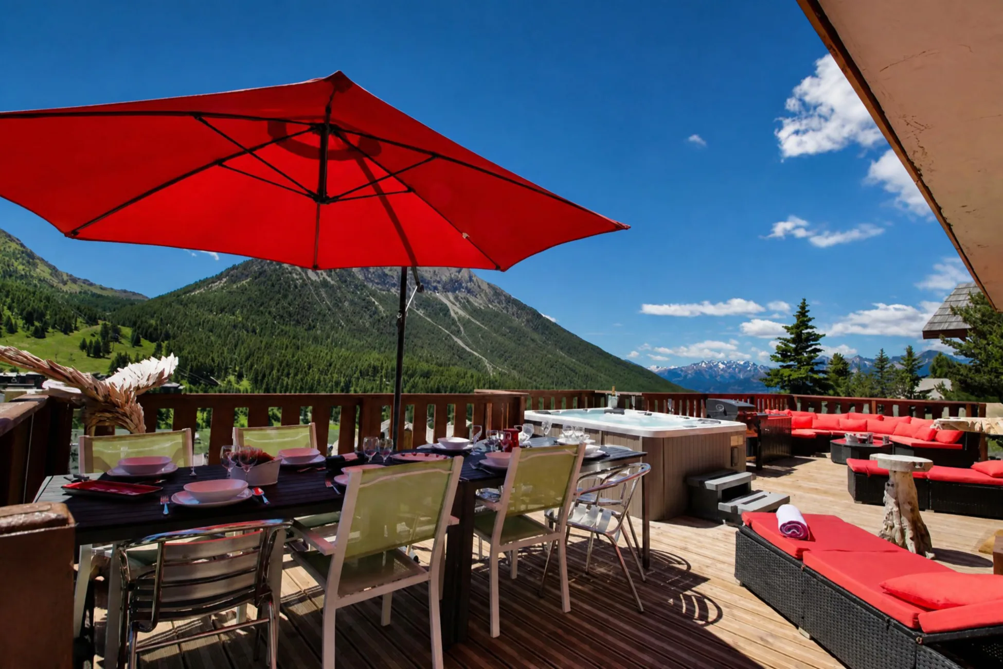 Grande terrasse ensoleillée du chalet La Soldanelle à Montgenèvre en été avec table dressée sous un parasol rouge, jacuzzi, barbecue et salon de jardin face aux montagnes