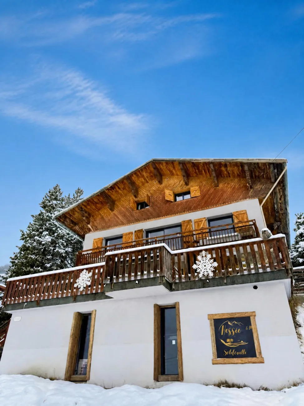 Façade du chalet La Soldanelle à Montgenèvre en hiver, avec balcons en bois, volets et sapins enneigés sous un ciel bleu